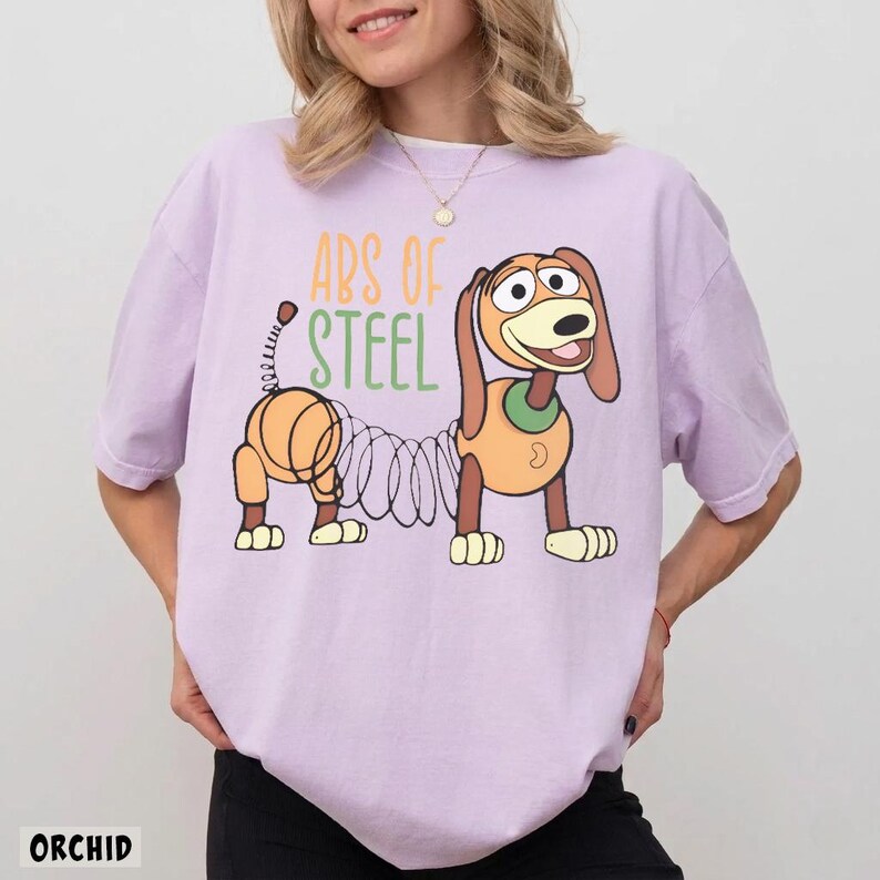 May include: A light purple t-shirt featuring a graphic of a cartoon dog with a spring-like body. The text "ABS OF STEEL" is printed above the dog in yellow and green lettering. The shirt has short sleeves and a crew neckline, made of cotton fabric.