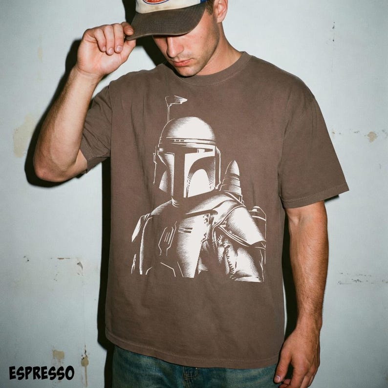 K&ouml;nnte beinhalten: Espressofarbenes T-Shirt mit einer detaillierten wei&szlig;en Grafik eines Mandalorianer-Helms und -R&uuml;stung. Das Shirt hat kurze &Auml;rmel und einen Rundhalsausschnitt. Das Wort "ESPRESSO" ist unten in Wei&szlig; aufgedruckt.