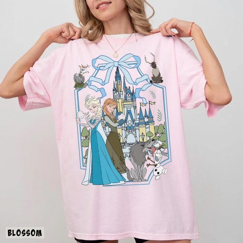 May include: A light pink t-shirt featuring a graphic of Elsa and Anna from Frozen, with a castle and other characters. The design is framed by a light blue bow and border. The word "BLOSSOM" is printed at the bottom.