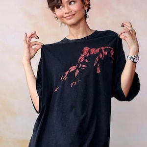 May include: A black short-sleeved t-shirt featuring a red graphic print of the superhero Spider-Man on the chest. The shirt has a crew neckline and a loose, relaxed fit. The graphic depicts the character in a crouched pose with web-like details.