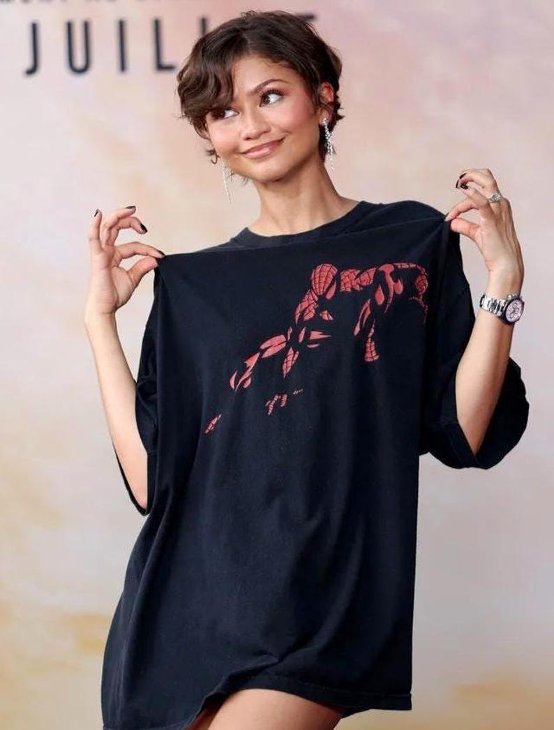 May include: A black short-sleeved t-shirt featuring a red graphic print of the superhero Spider-Man on the chest. The shirt has a crew neckline and a loose, relaxed fit. The graphic depicts the character in a crouched pose with web-like details.