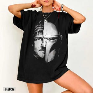 May include: Black t-shirt featuring a split image of a man's face and a Mandalorian helmet. The design is in black and white. The word "BLACK" is printed at the bottom of the shirt.
