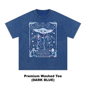 May include: Dark blue premium washed tee featuring a graphic of Grogu from Star Wars. The design includes the text "Grogu" and "Star Wars" in a decorative frame. The image is in a light blue and white color scheme.