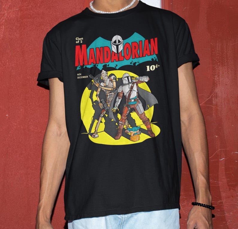 May include: Black t-shirt featuring a comic book style graphic of The Mandalorian and Grogu. The design includes the text "Clan of 2" and "MANDALORIAN" in red, with a 10&cent; price tag. The shirt has rolled-up sleeves.