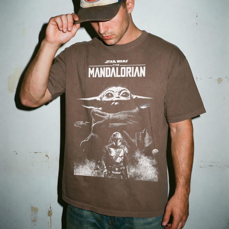 May include: Brown t-shirt featuring a graphic from the Star Wars series, "The Mandalorian." The design includes the text "STAR WARS THE MANDALORIAN" above an image of Grogu and the Mandalorian. The shirt is a solid brown color.