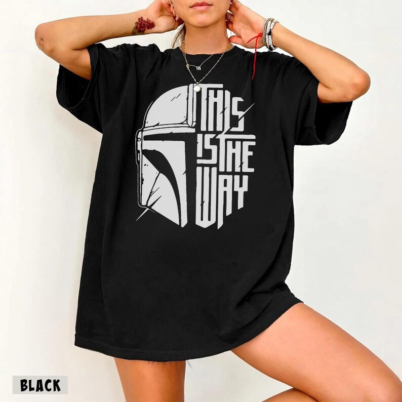 May include: Black t-shirt with a white graphic of a Mandalorian helmet and the text "THIS IS THE WAY". The shirt has a crew neck design.