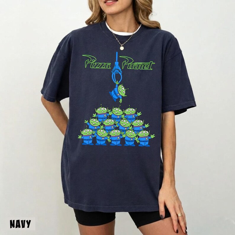 May include: Navy blue t-shirt featuring the Pizza Planet logo in green and a graphic of the Little Green Men from Toy Story. The design includes a green alien hanging from a claw and a group of aliens below.