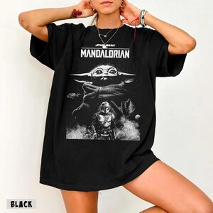 May include: Black t-shirt featuring a graphic from the Star Wars series, The Mandalorian. The design includes the text "STAR WARS MANDALORIAN" above an image of Grogu and the Mandalorian.