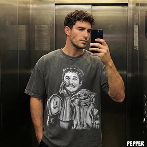 Puede incluir: Camiseta gris oscuro con un gr&aacute;fico en blanco y negro de un personaje con bigote y una peque&ntilde;a criatura verde. La camiseta tiene mangas cortas y cuello redondo. La imagen es un primer plano del dise&ntilde;o de la camiseta.