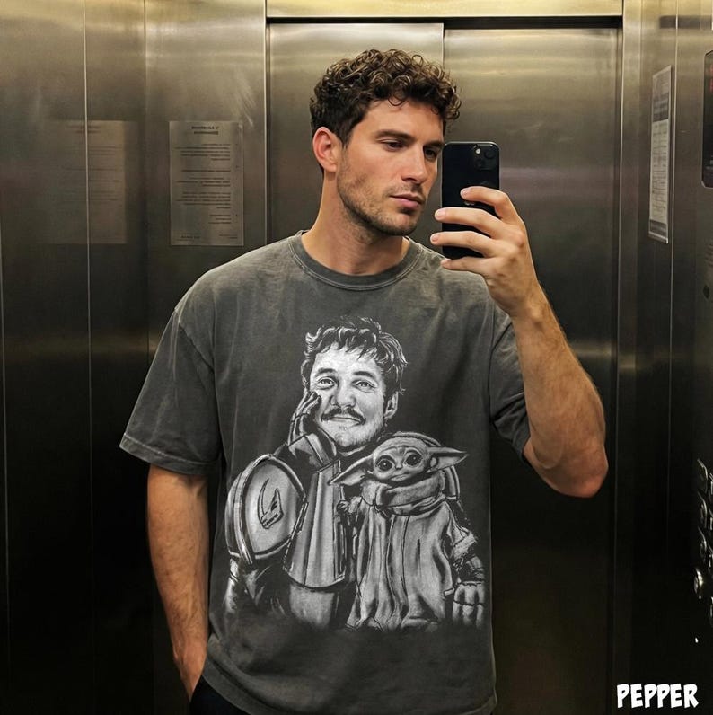 Puede incluir: Camiseta gris oscuro con un gr&aacute;fico en blanco y negro de un personaje con bigote y una peque&ntilde;a criatura verde. La camiseta tiene mangas cortas y cuello redondo. La imagen es un primer plano del dise&ntilde;o de la camiseta.