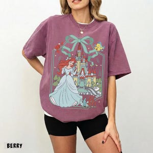 May include: Berry-colored t-shirt featuring a graphic of a princess in a blue gown, a castle, and aquatic characters. The design is framed by a teal bow and a decorative border. The word "BERRY" is printed at the bottom.