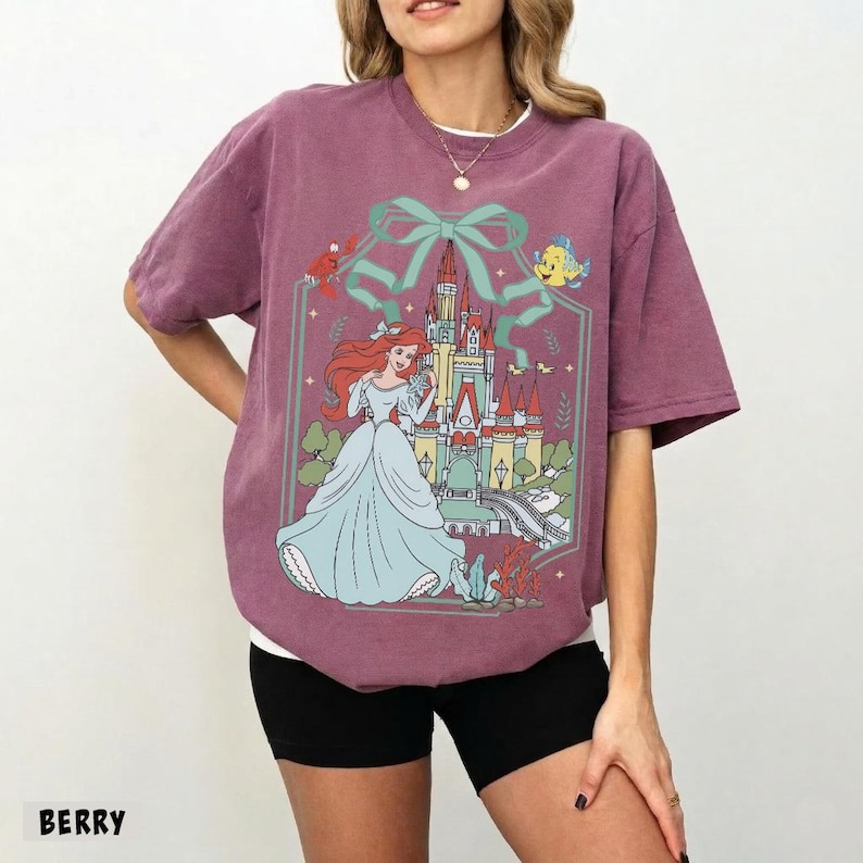 May include: Berry-colored t-shirt featuring a graphic of a princess in a blue gown, a castle, and aquatic characters. The design is framed by a teal bow and a decorative border. The word "BERRY" is printed at the bottom.