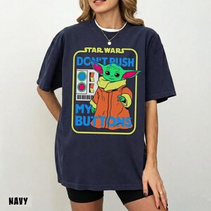 May include: Navy blue t-shirt featuring a graphic of Grogu from Star Wars. The design includes the text "STAR WARS DON'T PUSH MY BUTTONS" in a colorful, retro style. Grogu is depicted in his signature orange coat and green skin.
