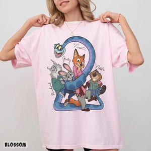 May include: A light pink t-shirt featuring a cartoon graphic of characters from Zootopia. The graphic includes Nick, Judy, and other characters, with the number 2 incorporated into the design. The word "BLOSSOM" is printed at the bottom.