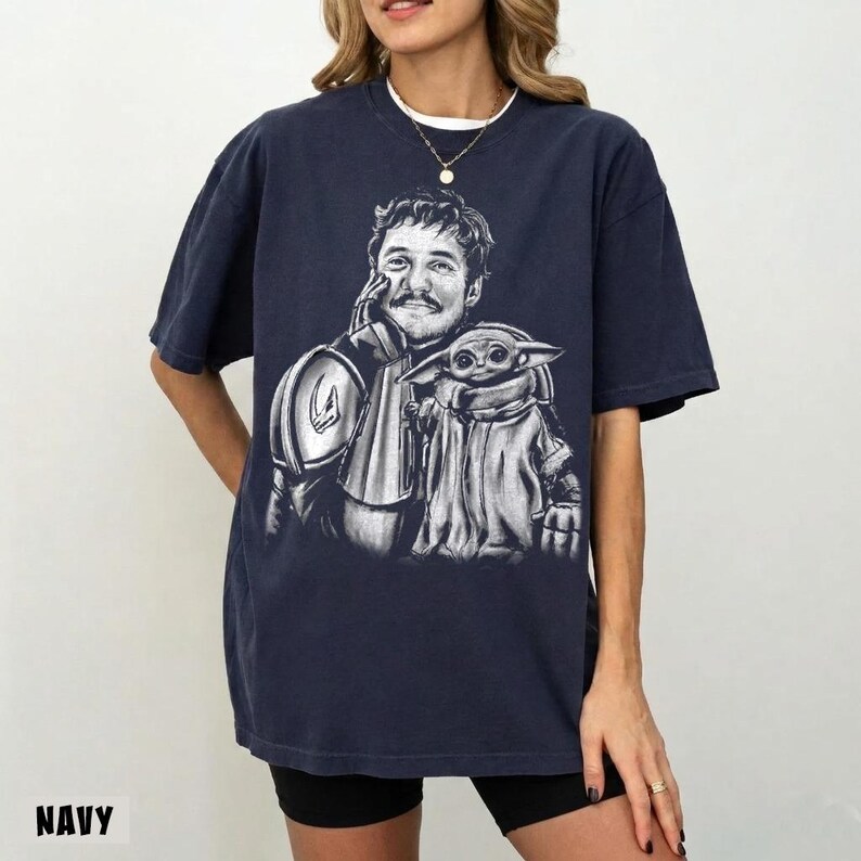 Puede incluir: Camiseta azul marino con un gr&aacute;fico en blanco y negro de un personaje con bigote y un peque&ntilde;o alien&iacute;gena verde. La palabra "NAVY" est&aacute; impresa en la parte inferior. La camiseta tiene mangas cortas y un corte holgado.