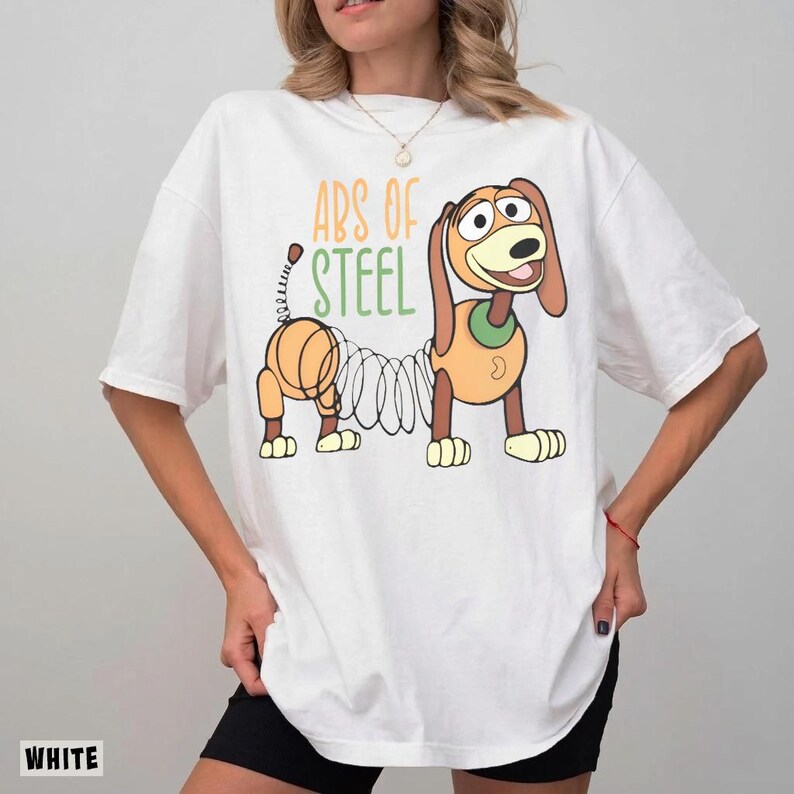 May include: A white short-sleeved t-shirt featuring a graphic of a brown and tan toy dog with a coiled spring body. Above the dog, the text reads "ABS OF STEEL" in orange and green letters. The shirt has a crew neckline and a relaxed fit.