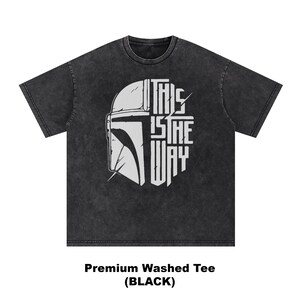 May include: Black washed tee with a white graphic of a helmet and the text "THIS IS THE WAY". The design is centered on the front of the shirt. The shirt is a premium washed tee.
