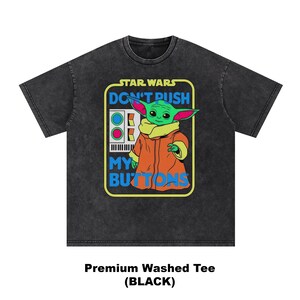 May include: Black washed tee featuring a graphic of Grogu from Star Wars. The design includes the text "DON'T PUSH MY BUTTONS" and a control panel. Grogu is depicted in his orange coat and tan collar. The shirt is a premium washed tee.