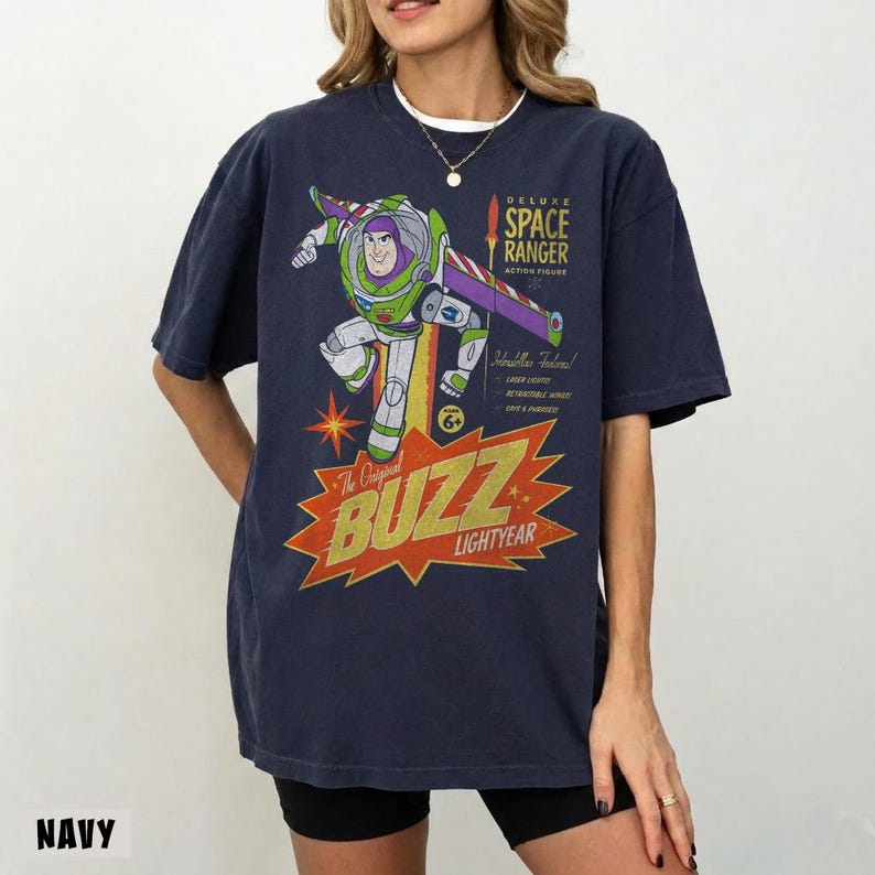 May include: A navy blue short-sleeve t-shirt featuring a graphic of the Buzz Lightyear character in a space suit. The design includes the text Deluxe Space Ranger Action Figure and Buzz Lightyear in a large, stylized orange and yellow font.