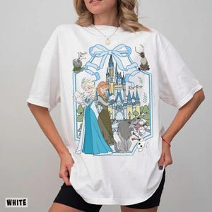 May include: White t-shirt featuring a cartoon graphic of Elsa and Anna from Frozen, with a castle and other characters. The design is framed by a light blue ribbon and a hexagonal border. The word "WHITE" is printed at the bottom.