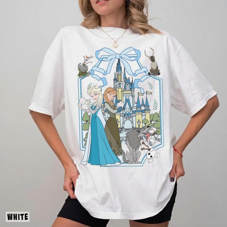 May include: White t-shirt featuring a cartoon graphic of Elsa and Anna from Frozen, with a castle and other characters. The design is framed by a light blue ribbon and a hexagonal border. The word "WHITE" is printed at the bottom.