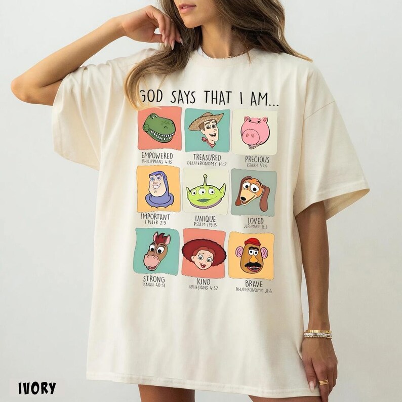 May include: Ivory-colored t-shirt with the text "GOD SAYS THAT I AM..." and colorful cartoon character graphics. Each character is in a square with a descriptive word and a Bible verse reference. The word "IVORY" is printed at the bottom.
