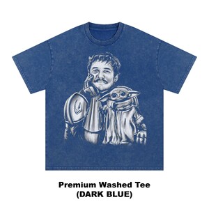 Puede incluir: Camiseta lavada premium azul oscuro con un gr&aacute;fico de un hombre y una peque&ntilde;a criatura. El hombre lleva una armadura y la criatura est&aacute; envuelta en una capa. El texto "Premium Washed Tee (DARK BLUE)" est&aacute; en la parte inferior.