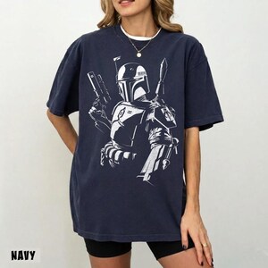 May include: Navy blue t-shirt featuring a white graphic of a character with a helmet, holding a blaster and a weapon. The word "NAVY" is printed at the bottom of the shirt. The shirt has short sleeves and a relaxed fit.