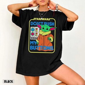 May include: Black t-shirt featuring a graphic of Grogu from Star Wars with the text "DON'T PUSH MY BUTTONS." The design includes a green Grogu wearing an orange coat and a yellow collar. The Star Wars logo is at the top.