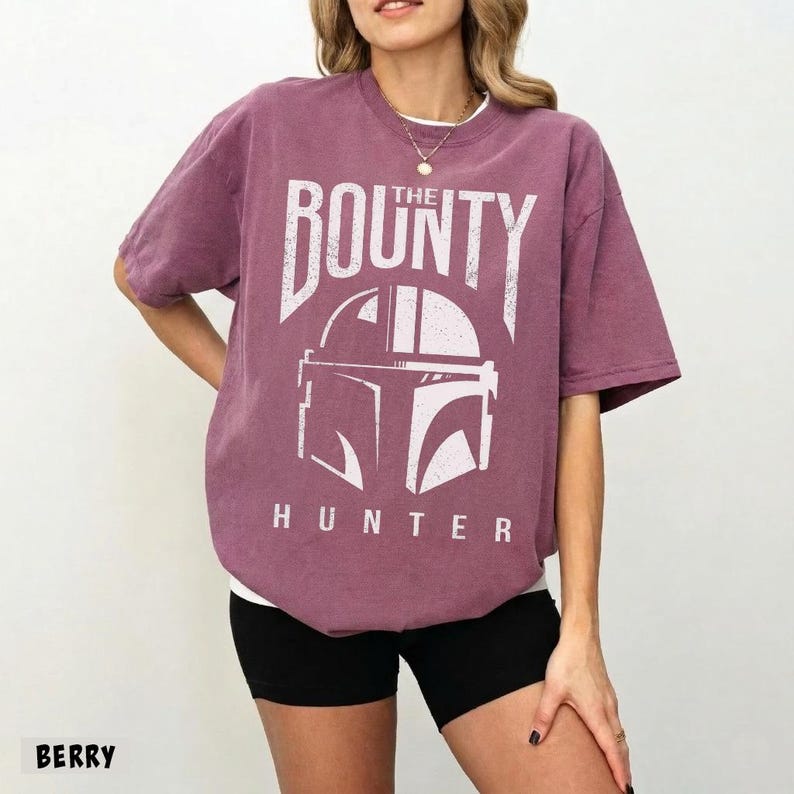 Puede incluir: Camiseta color bayas con el texto "THE BOUNTY HUNTER" y un gr&aacute;fico blanco de un casco. La camiseta es de corte holgado y tiene mangas cortas. La modelo lleva un pantal&oacute;n corto negro.