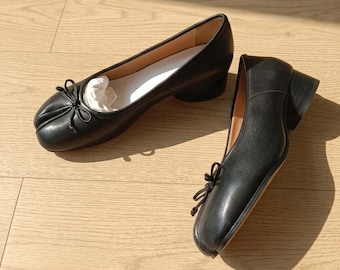 May include: A pair of black leather shoes with a split-toe design, featuring a small decorative bow on the front, a low block heel, and a light-coloured interior lining. The shoes have a smooth finish and a structured silhouette with a rounded toe.