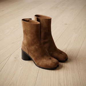 May include: A pair of brown suede ankle boots featuring a distinct split-toe design, a mid-height cylindrical black block heel, and a smooth tan interior lining. The boots have a clean, structured silhouette with a side seam detail.