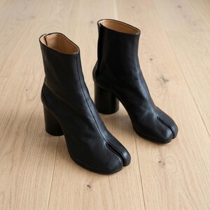 May include: A pair of black leather ankle boots featuring a distinct split-toe design, a cylindrical block heel, and a smooth finish. The boots have a high shaft with a vertical seam running down the front and a tan interior lining visible at the top.
