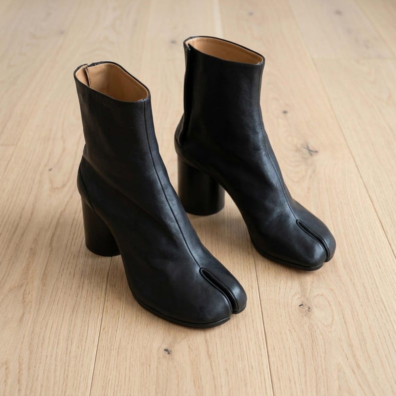 May include: A pair of black leather ankle boots featuring a distinct split-toe design, a cylindrical block heel, and a smooth finish. The boots have a high shaft with a vertical seam running down the front and a tan interior lining visible at the top.