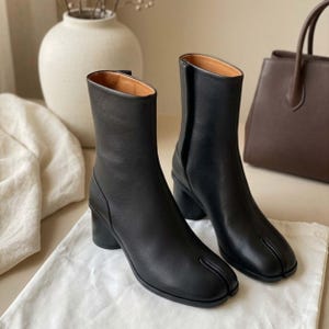 May include: A pair of black leather ankle boots featuring a distinct split-toe design, a mid-height cylindrical block heel, and a vertical seam running up the front of the shaft. The boots have a smooth finish and a tan interior lining.