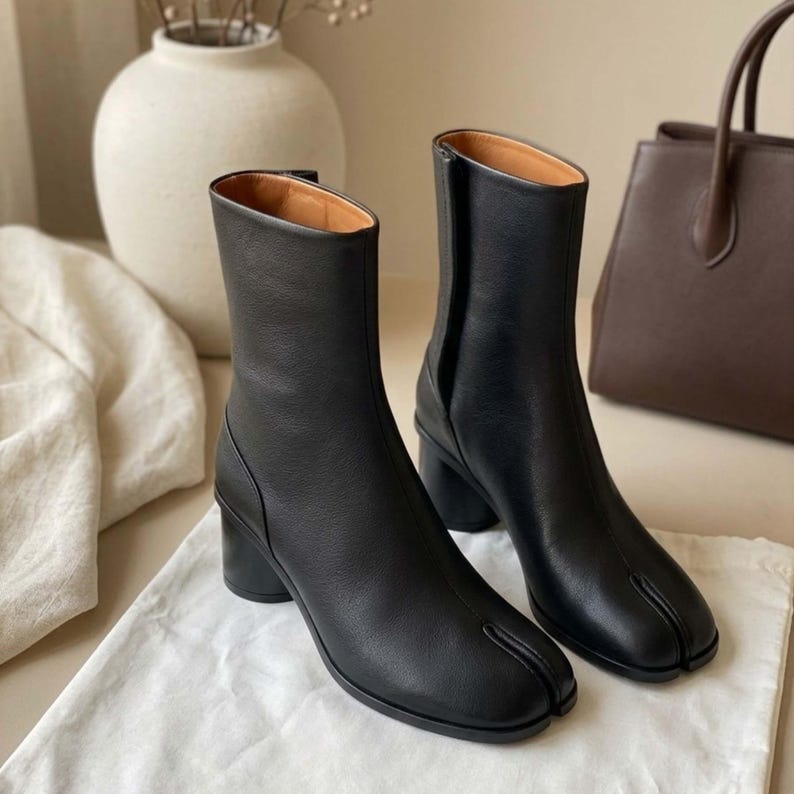 May include: A pair of black leather ankle boots featuring a distinct split-toe design, a mid-height cylindrical block heel, and a vertical seam running up the front of the shaft. The boots have a smooth finish and a tan interior lining.