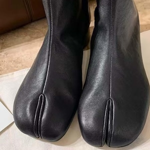 May include: A pair of black leather ankle boots featuring a distinct split-toe design. The boots have a smooth, matte finish with a vertical seam running up the center of the vamp, separating the big toe from the rest of the foot area.