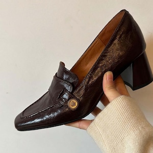 May include: A dark brown, crinkled-texture leather loafer pump with a square toe and a block heel. The shoe features a decorative gold-toned circular emblem with a floral motif on the side of the vamp, and a tan leather interior lining.