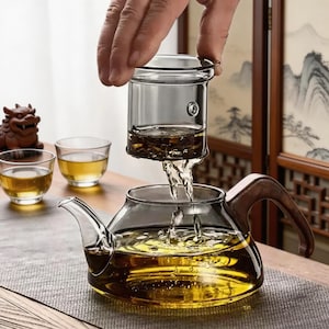 May include: A clear glass teapot with a wooden handle is being filled with tea from a glass infuser. Two small glass teacups and a decorative statue are on the table. The tea is a golden yellow color.
