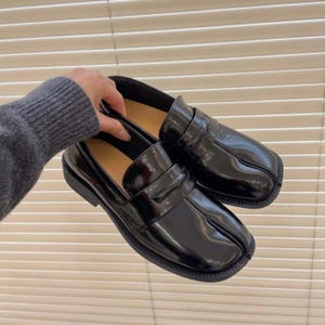Pode incluir: Um par de mocassins pretos com acabamento brilhante e um design distinto de biqueira dividida. Estes sapatos apresentam uma tira clássica de mocassim sobre o peito do pé, uma forma de biqueira quadrada e uma sola de borracha preta texturizada.