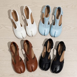 May include: Four pairs of split-toe shoes arranged in two rows. The top row features white and light blue shoes, while the bottom row displays brown and black shoes. Each shoe has a distinct T-strap design and a separated toe compartment.