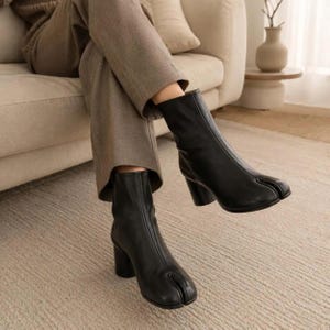 May include: A pair of black ankle boots featuring a distinct split-toe design, a smooth finish, and a sturdy block heel. The boots extend to the mid-calf and are worn with neutral-toned trousers, showcasing a clean, structured silhouette.