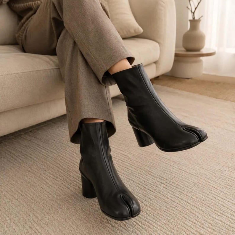 May include: A pair of black ankle boots featuring a distinct split-toe design, a smooth finish, and a sturdy block heel. The boots extend to the mid-calf and are worn with neutral-toned trousers, showcasing a clean, structured silhouette.