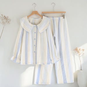 Könnte beinhalten: Ein zweiteiliger Pyjama-Set mit Streifen in Hellblau, Creme und Weiß. Das langärmlige Oberteil hat einen großen, runden Kragen und eine Knopfleiste. Die Hose ist locker geschnitten und hat einen elastischen Bund. Das Set hängt an einem Holzbügel.