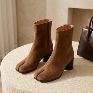 May include: A pair of brown suede ankle boots featuring a split-toe design, a mid-height cylindrical black heel, and a side zipper closure. The boots have a smooth, matte texture and a structured silhouette that reaches above the ankle.