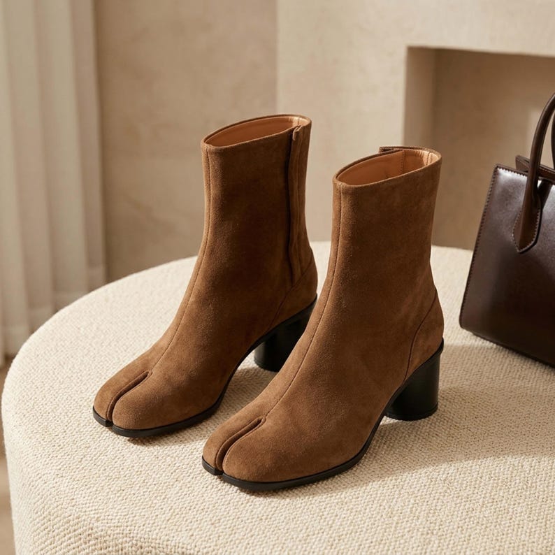 May include: A pair of brown suede ankle boots featuring a split-toe design, a mid-height cylindrical black heel, and a side zipper closure. The boots have a smooth, matte texture and a structured silhouette that reaches above the ankle.