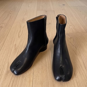 May include: A pair of black ankle boots featuring a distinct split-toe design. The boots have a smooth finish, a vertical seam running up the front, and a low cylindrical heel. The interior lining is a light tan color, and the boots are ankle-high.