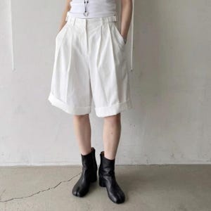 May include: White knee-length shorts featuring a high-waisted design with front pleats, a button and zipper closure, belt loops, and wide cuffed hems. The shorts are paired with black leather ankle boots that have a split-toe design.