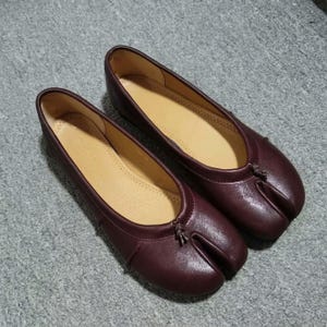 May include: A pair of dark burgundy leather ballet flats featuring a split-toe design. The shoes have a smooth, matte finish with a small knotted detail at the center of the vamp and a light tan interior lining, showcasing a low-profile silhouette.