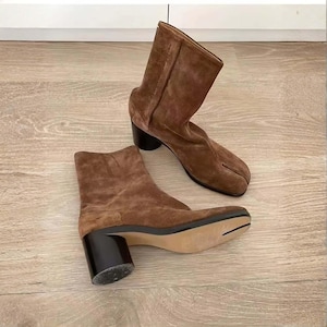 May include: A pair of brown suede ankle boots featuring a distinct split-toe design. The boots have a mid-height cylindrical black heel and a smooth, light-colored sole. The suede material has a soft, textured appearance with visible vertical seams.